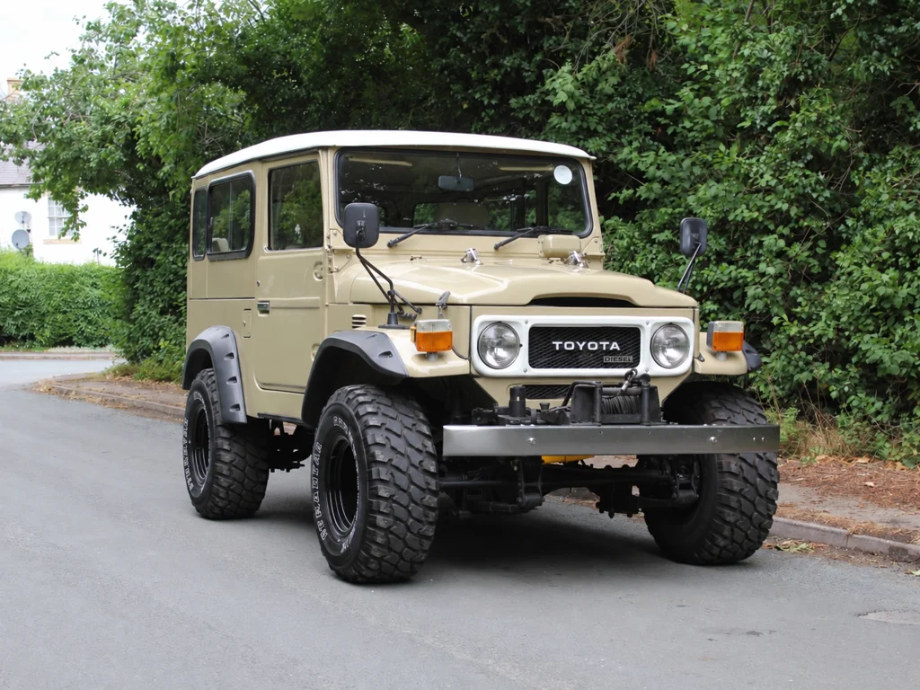 Beige classic Land Cruiser with a white roof, parked beside green hedges